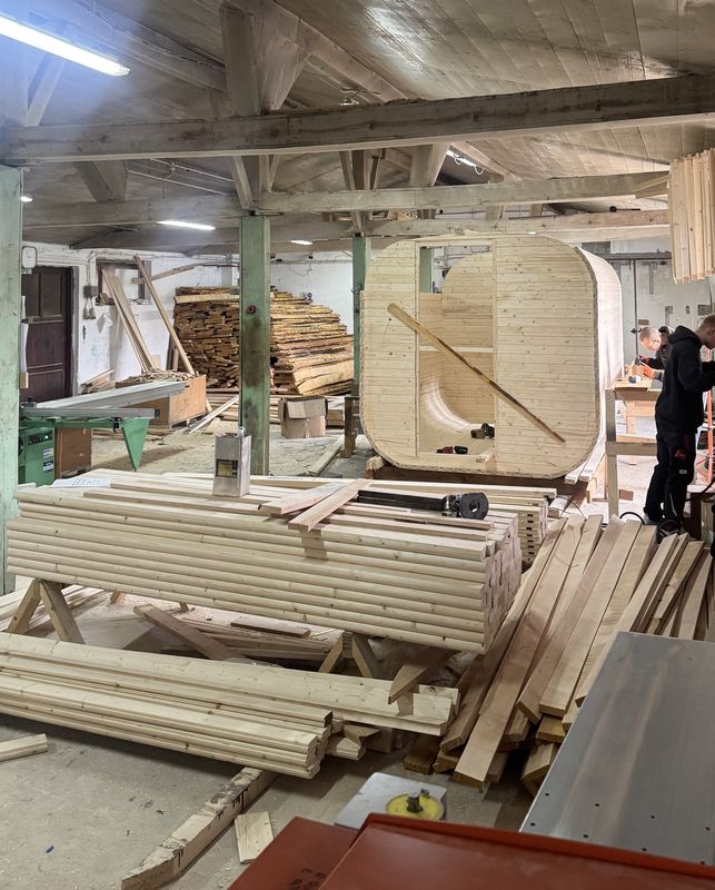 Hala produkcyjna WarmEscape — stosy desek świerkowych, w głębi sauna Quadro w budowie