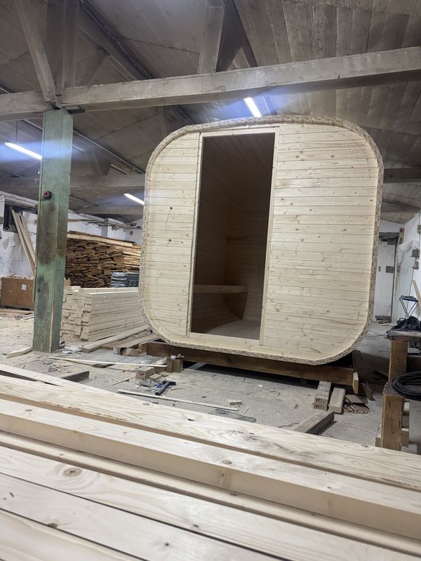 Surowa sauna Quadro ze świerku w hali WarmEscape, w tle stosy desek